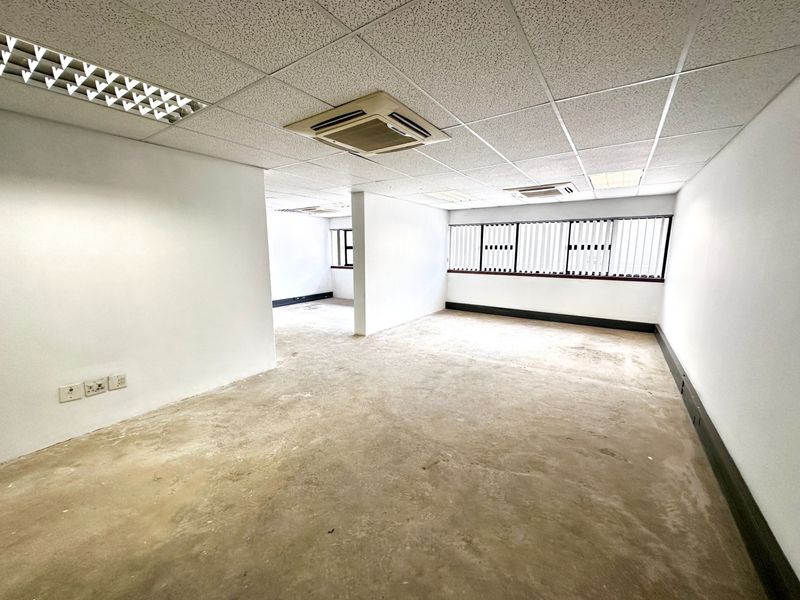 An empty, open-plan office space featuring a suspended ceiling, recessed lighting, ceiling-mounted air conditioning units, and large windows with horizontal blinds.