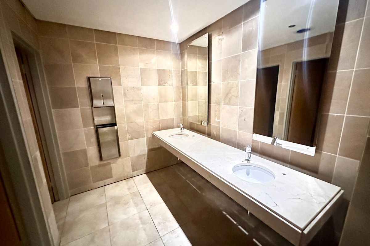 A clean, modern office bathroom featuring a double vanity with stone countertops, undermount sinks, large wall-mounted mirrors, and neutral floor-to-ceiling tiling.