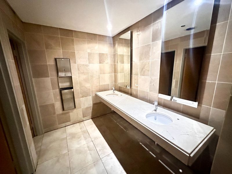 A clean, modern office bathroom featuring a double vanity with stone countertops, undermount sinks, large wall-mounted mirrors, and neutral floor-to-ceiling tiling.