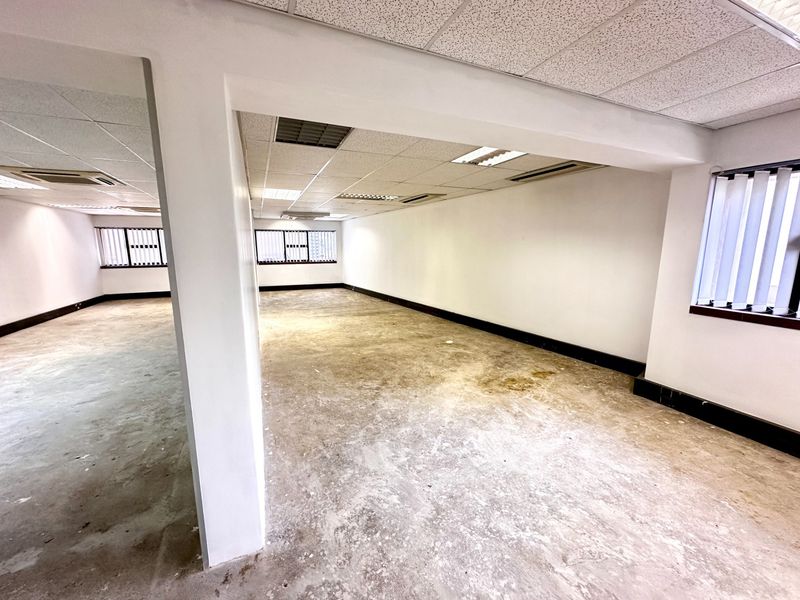 An empty, open-plan office space featuring a suspended ceiling, recessed lighting, and vertical blinds, requiring flooring refurbishment.