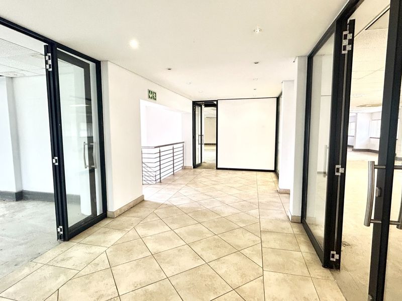 A spacious, vacant office hallway featuring tiled flooring, modern glass partition doors, and access to a staircase, offering a clean and professional layout.