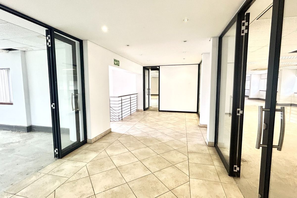 A spacious, vacant office hallway featuring tiled flooring, modern glass partition doors, and access to a staircase, offering a clean and professional layout.