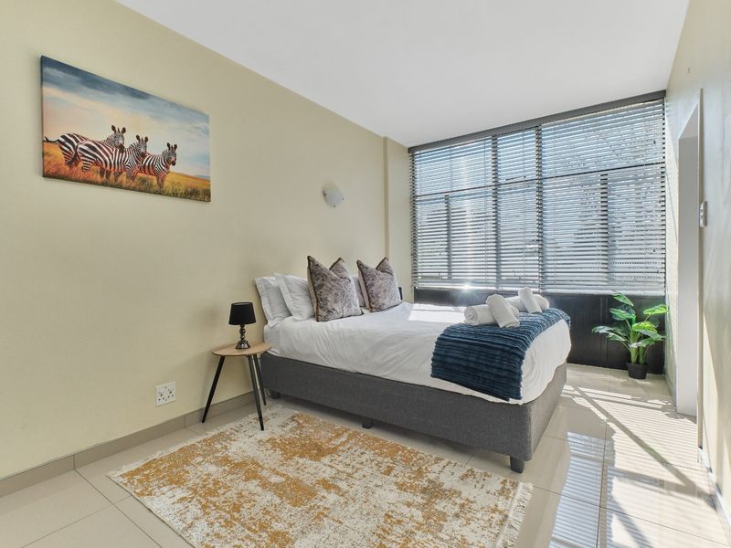 A well-lit bedroom featuring neutral tiled flooring, large windows with venetian blinds, and a wall-mounted light fixture.
