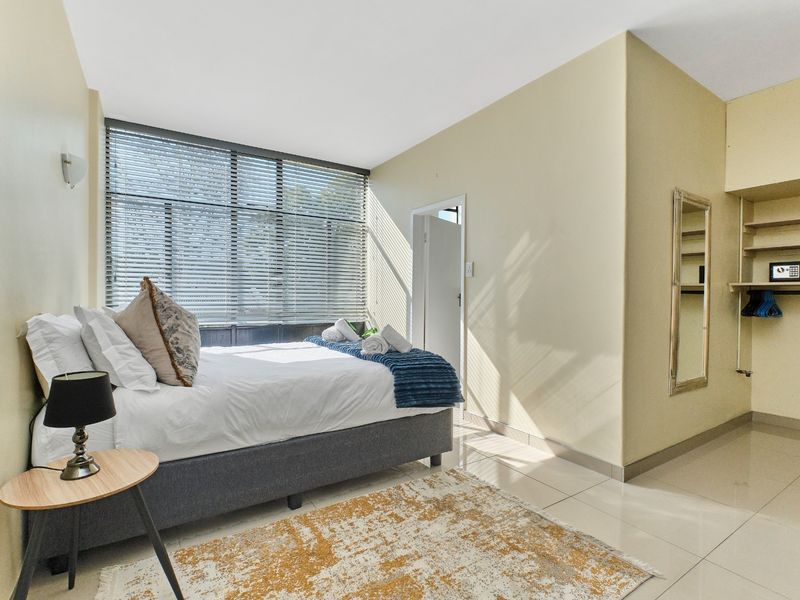 A bright bedroom featuring large windows with venetian blinds, tiled flooring, and built-in open shelving for storage.