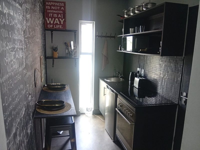 Compact kitchen featuring dark cabinetry, built-in oven, electric hob, stainless steel sink, and a tiled backsplash with a narrow window for natural light.