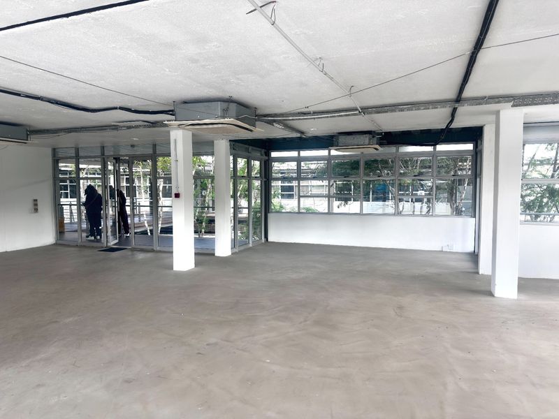 An open-plan office space featuring concrete floors, exposed ceiling infrastructure, ducted air conditioning, and large windows providing ample natural light.