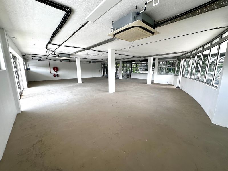 A spacious, open-plan office area featuring exposed ceiling services, structural columns, and large windows providing ample natural light.