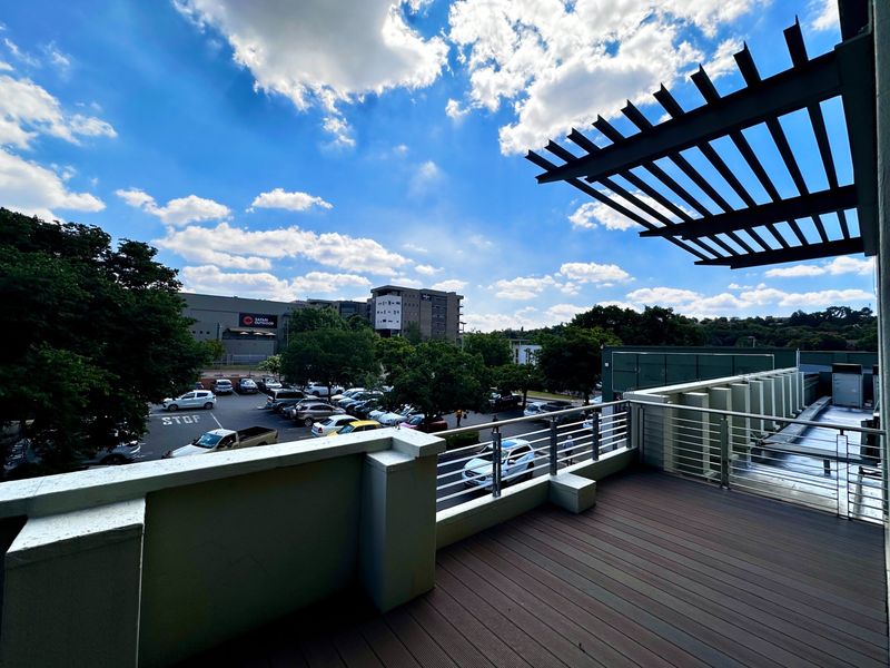 A spacious outdoor balcony featuring timber decking, a modern metal balustrade, and an overhead pergola, offering an elevated view of the surrounding parking area.