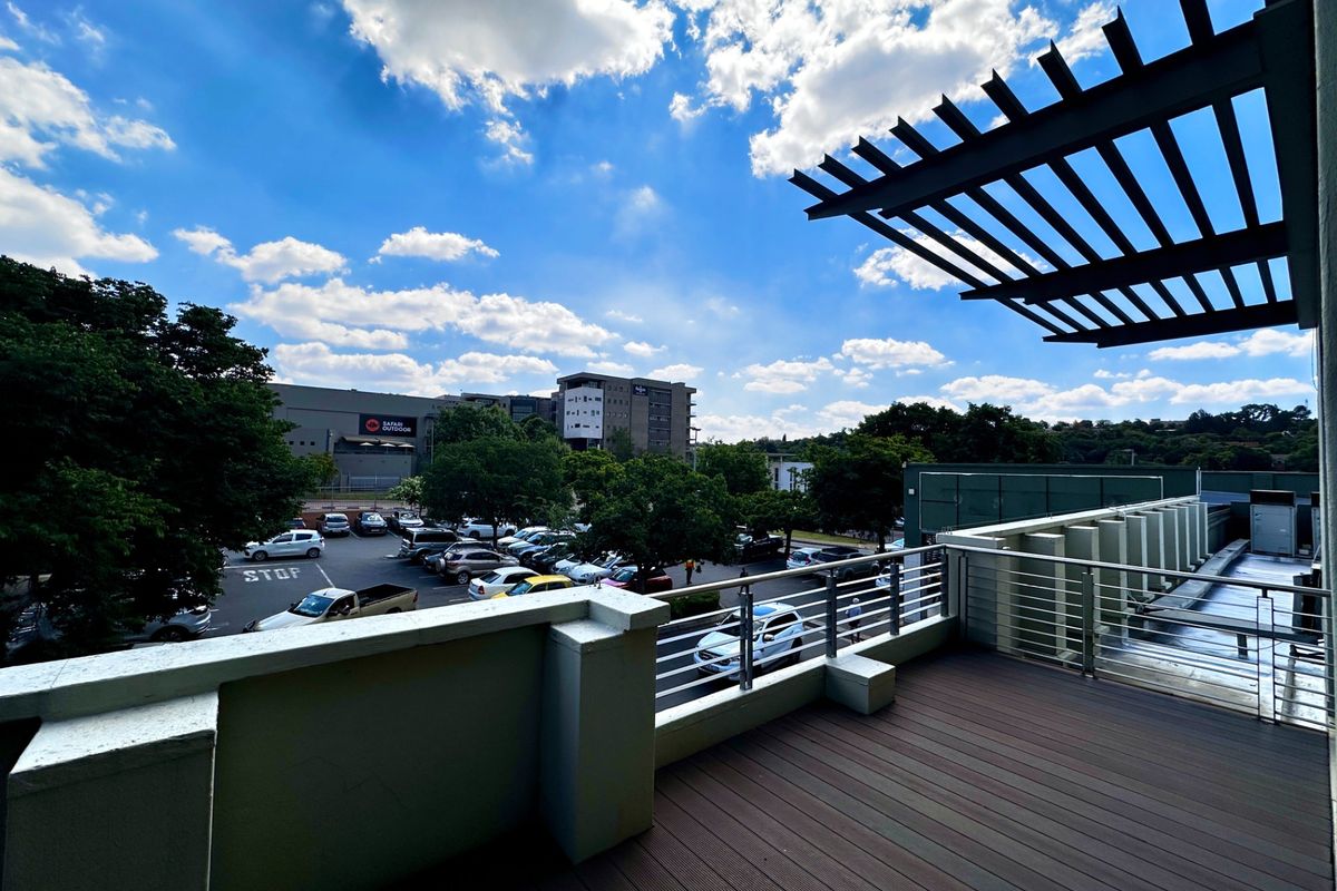 A spacious outdoor balcony featuring timber decking, a modern metal balustrade, and an overhead pergola, offering an elevated view of the surrounding parking area.