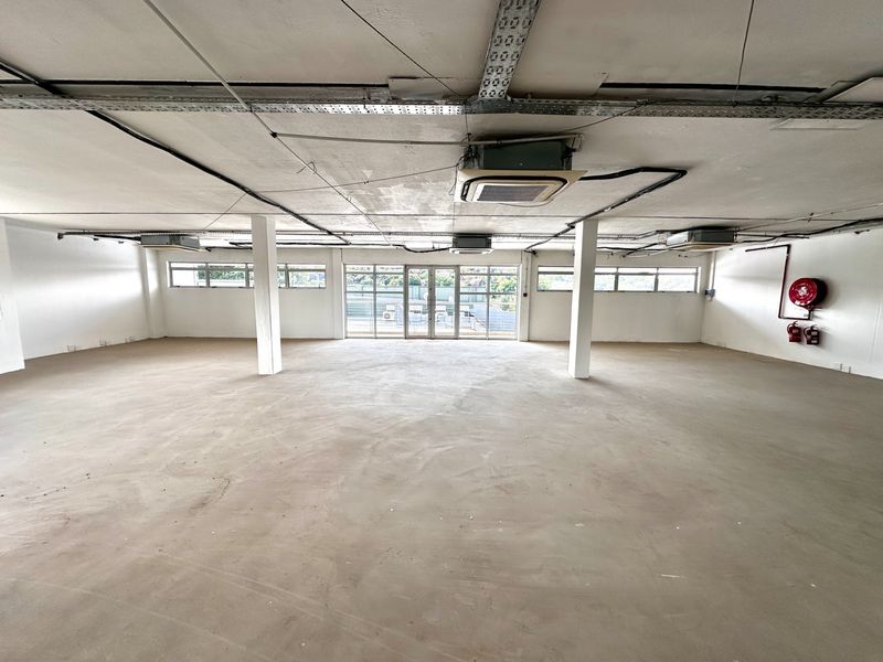 Spacious open-plan office area featuring exposed industrial ceilings, cassette air conditioning, and large windows providing natural light and access to a balcony.