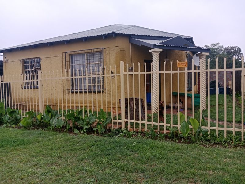 Front view of a single-story house featuring a corrugated metal roof, decorative columns, and perimeter security fencing.