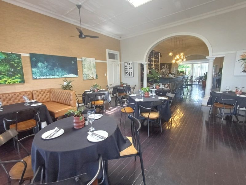 Spacious open-plan dining area featuring dark hardwood flooring, elegant crown molding, a ceiling fan, and a distinct arched transition to a built-in bar area.