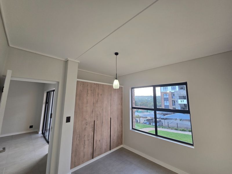 A bright, modern bedroom featuring built-in wood-finish cupboards, neutral tiled flooring, and large aluminium-framed windows with a pleasant exterior view.