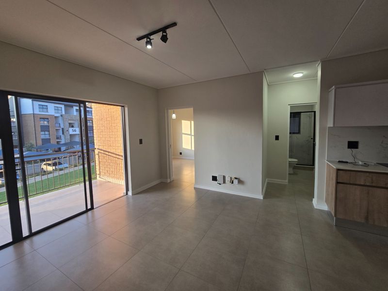Modern open-plan living area featuring neutral tiled flooring, track lighting, and seamless access to a private balcony, adjacent to a contemporary kitchen.