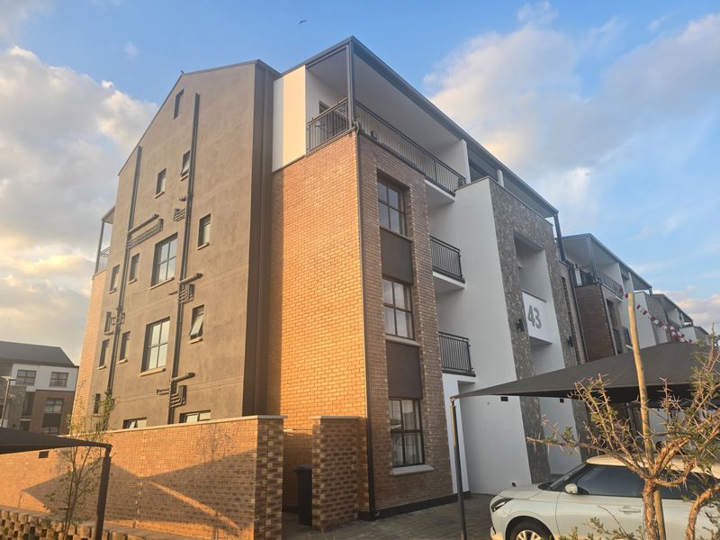 A modern multi-story apartment building featuring a combination of brick and rendered finishes, private balconies, and a paved parking area with shade structures.