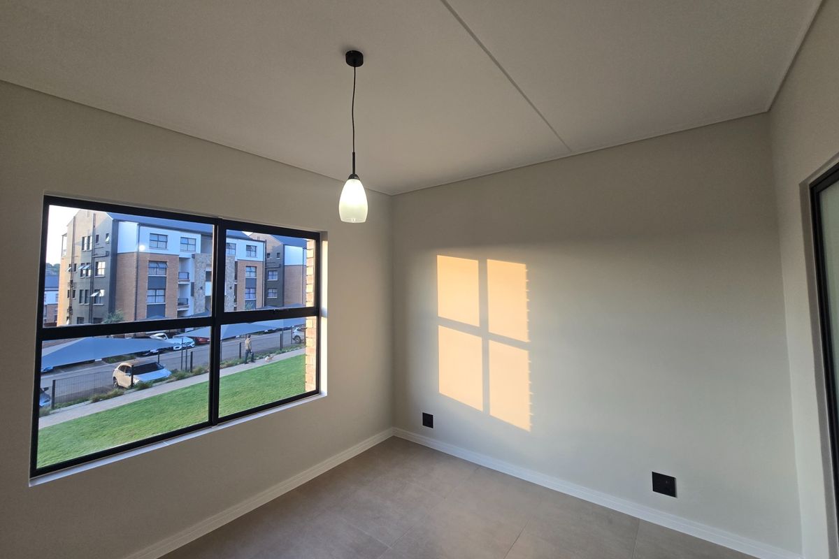A bright, empty bedroom featuring modern tiled flooring, a pendant light fixture, and large aluminium-framed windows with a view of the complex.