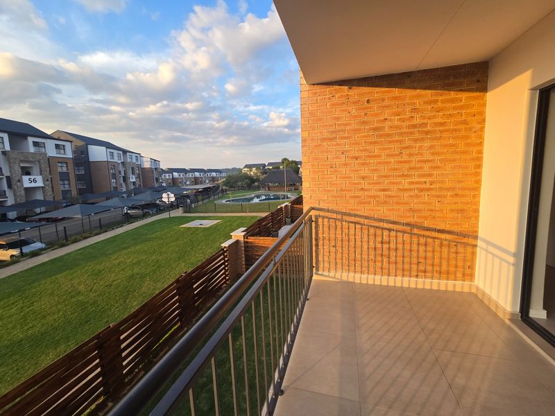 A clean, modern balcony featuring tiled flooring, a metal balustrade, and an exposed brick wall, overlooking the complex grounds and pool area.