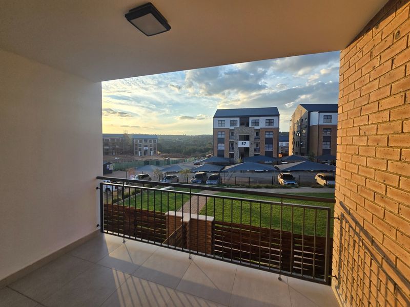 A clean, covered balcony featuring tiled flooring and a metal balustrade, offering an elevated view of the apartment complex grounds and surrounding area.