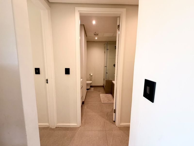 A clean, neutral hallway featuring tiled flooring and recessed lighting, providing access to an adjacent bathroom with a glass-enclosed shower.