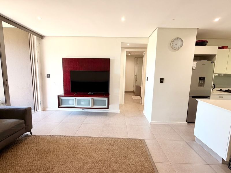 Modern open-plan living area featuring tiled floors, recessed lighting, and a built-in media unit, seamlessly connecting to the kitchen and balcony access.