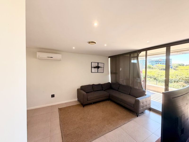A bright living room featuring tiled flooring, recessed downlighting, a wall-mounted air conditioning unit, and large sliding glass doors leading to a balcony.