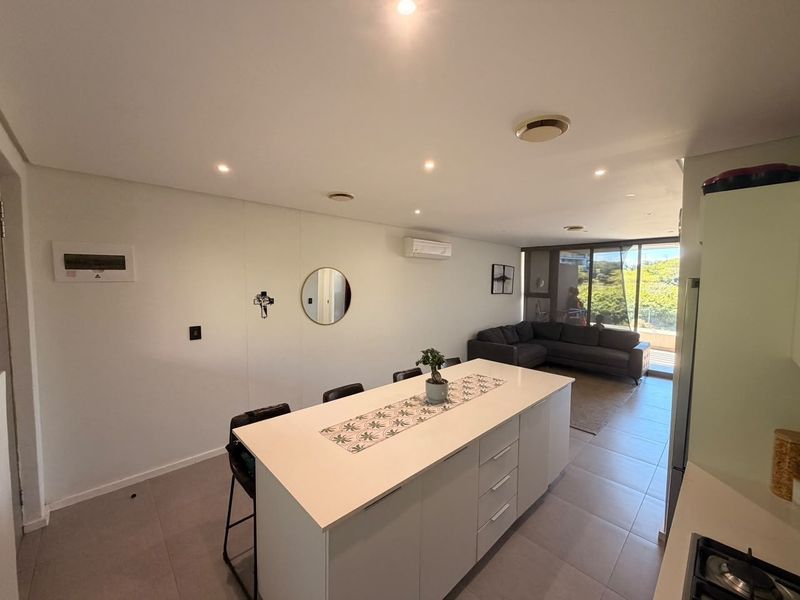 Modern open-plan living area featuring a central kitchen island, tiled flooring, recessed lighting, and seamless access to an outdoor balcony through sliding glass doors.