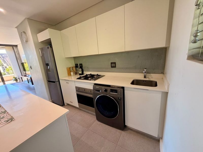 Modern kitchen featuring sleek white cabinetry, stone countertops, a gas hob, built-in oven, and integrated laundry space, leading to an open-plan living area.