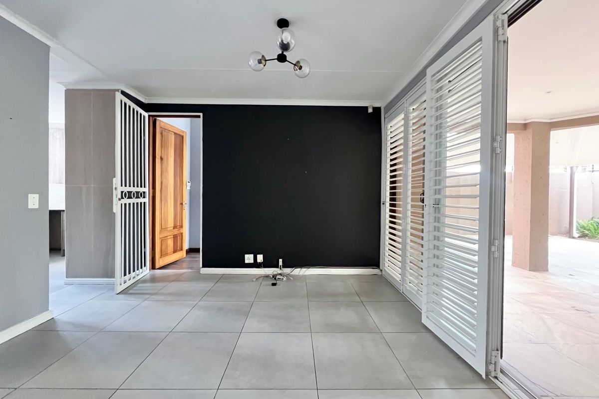 A bright, open-plan living area featuring large format floor tiles, a striking black accent wall, and sliding shutters leading to an outdoor patio.