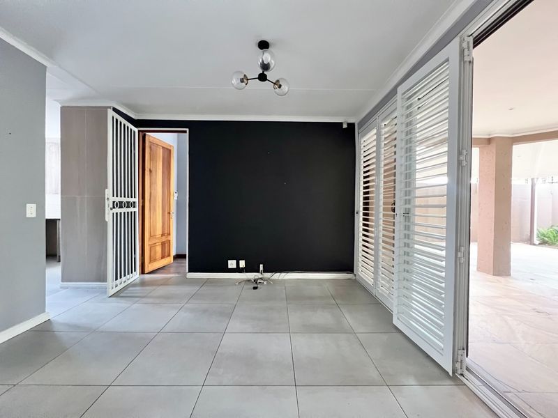 A bright, open-plan living area featuring large format floor tiles, a striking black accent wall, and sliding shutters leading to an outdoor patio.