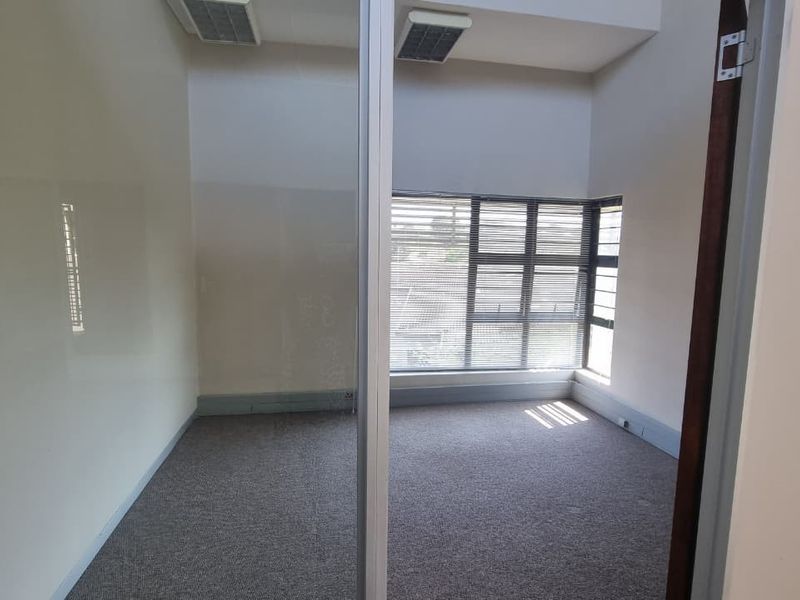 A bright, empty office space featuring carpeted flooring, a large window with blinds, and a modern aluminum-framed glass partition.