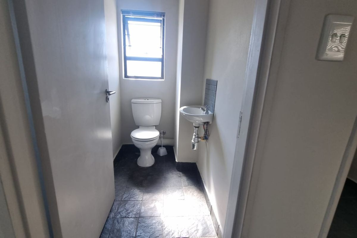 A compact office bathroom featuring a toilet, wall-mounted basin, and dark tiled flooring with natural light from a top-hung window.