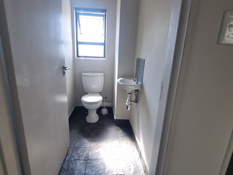 A compact office bathroom featuring a toilet, wall-mounted basin, and dark tiled flooring with natural light from a top-hung window.