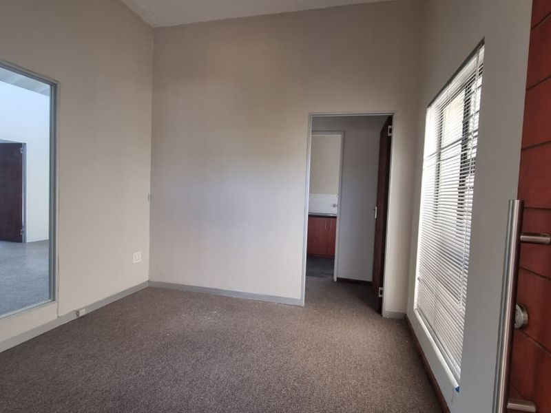 A bright, empty office space featuring carpeted flooring, a large glass partition, and a solid wood entrance door with integrated venetian blinds.