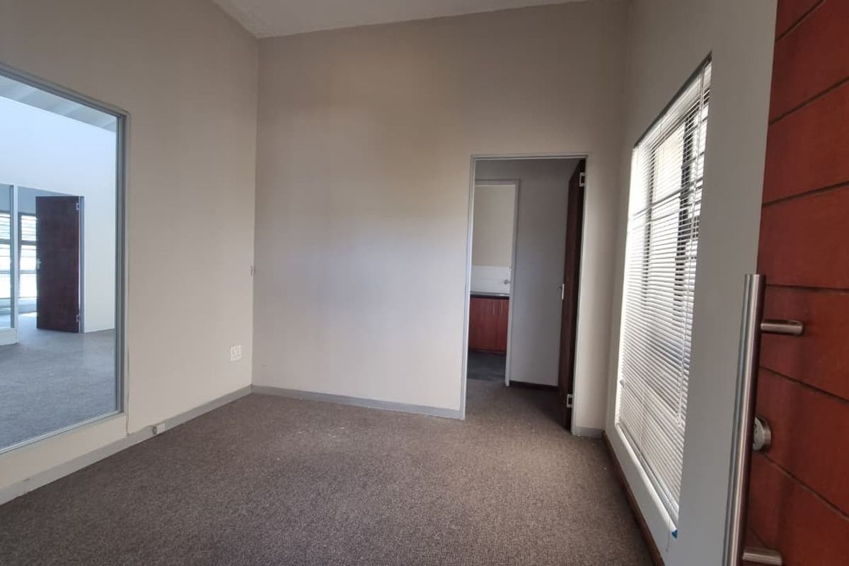 A bright, empty office space featuring carpeted flooring, a large glass partition, and a solid wood entrance door with integrated venetian blinds.