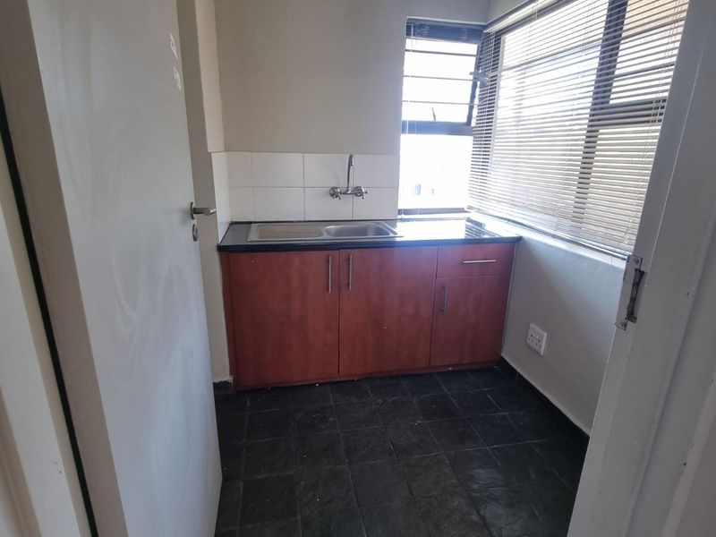A compact kitchenette featuring dark cabinetry, a stainless steel sink, tiled backsplash, and large windows with venetian blinds.