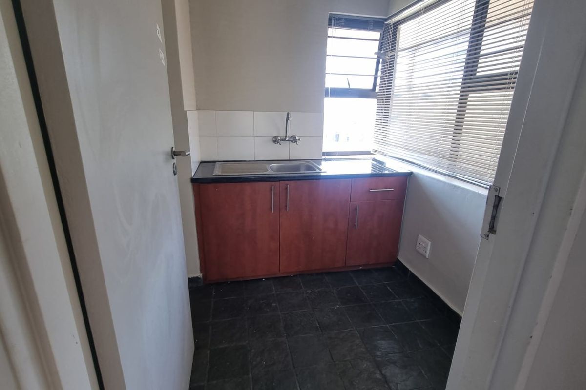 A compact kitchenette featuring dark cabinetry, a stainless steel sink, tiled backsplash, and large windows with venetian blinds.