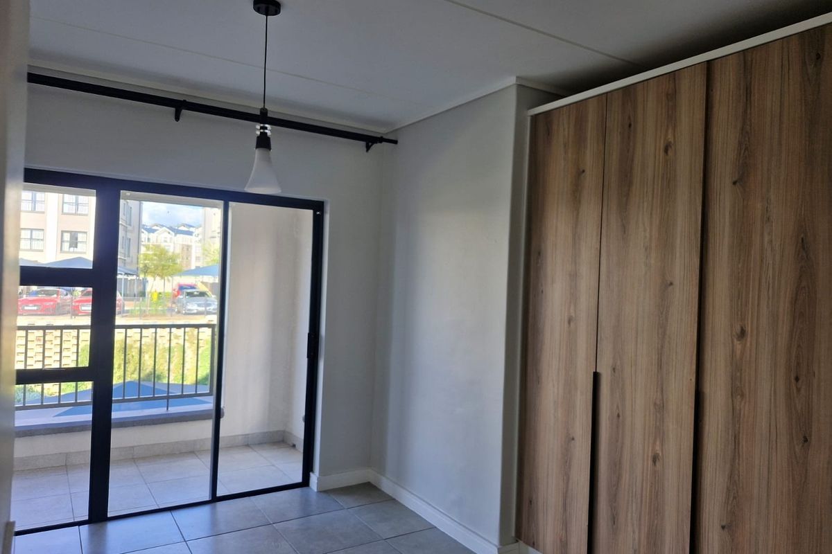 A bright, empty bedroom featuring modern wood-finish built-in cupboards, tiled flooring, and a sliding glass door leading to a private balcony.