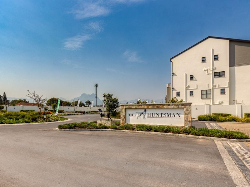 The entrance to The Huntsman apartment complex, featuring a paved driveway, landscaped gardens, and secure boundary walling with electric fencing.