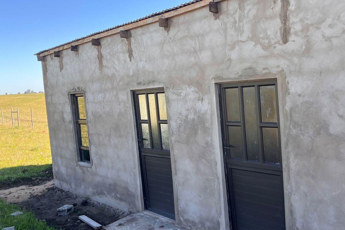 Exterior view of a small holding structure featuring raw cement plaster walls, corrugated iron roofing, and dark aluminium framed doors and windows.