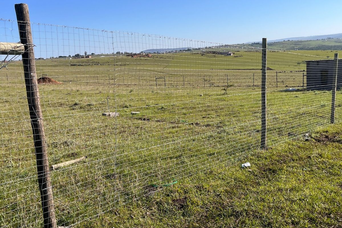 Expansive open land featuring wire mesh perimeter fencing and a small utility structure, set against a rural landscape backdrop.