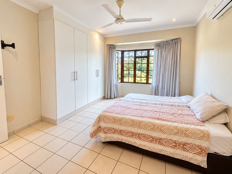 A well-maintained bedroom featuring built-in cupboards, a ceiling fan, air conditioning, and a bay window with natural light.