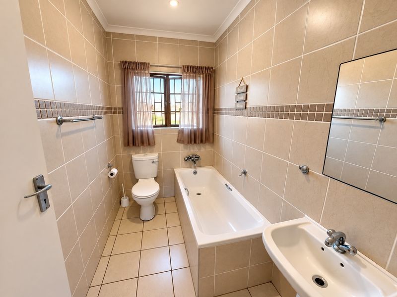 A clean, functional bathroom featuring neutral wall and floor tiles, a standard bathtub, pedestal basin, and a window for natural light.