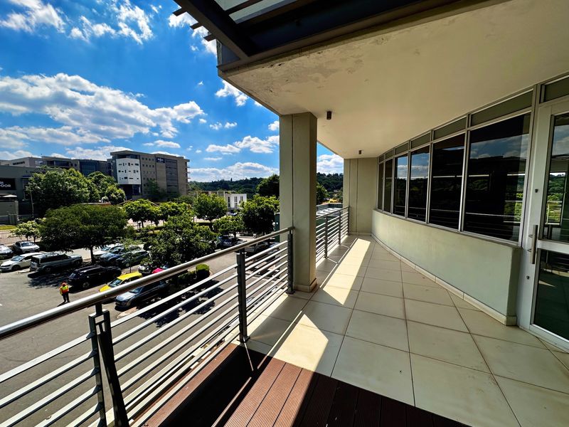 A spacious office balcony featuring tiled flooring, timber decking, and a metal balustrade, offering an elevated view of the surrounding parking area and office park.