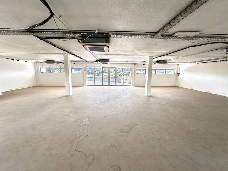 A spacious, open-plan office area featuring exposed ceiling services, concrete flooring, and large windows providing natural light and exterior views.