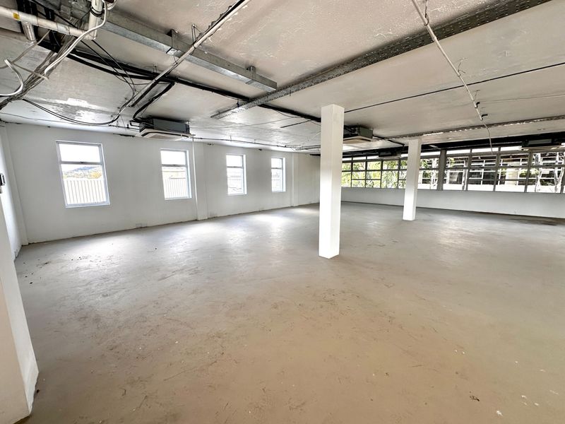 Open-plan office space featuring exposed industrial ceilings, structural columns, and large windows providing ample natural light.