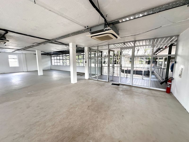 Spacious open-plan office area featuring exposed ceiling infrastructure, concrete flooring, and large glass partitions leading to an outdoor balcony.