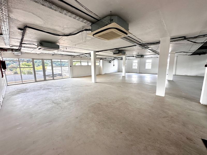 Spacious open-plan office area featuring exposed industrial ceilings, structural columns, concrete flooring, and large glass doors providing natural light.