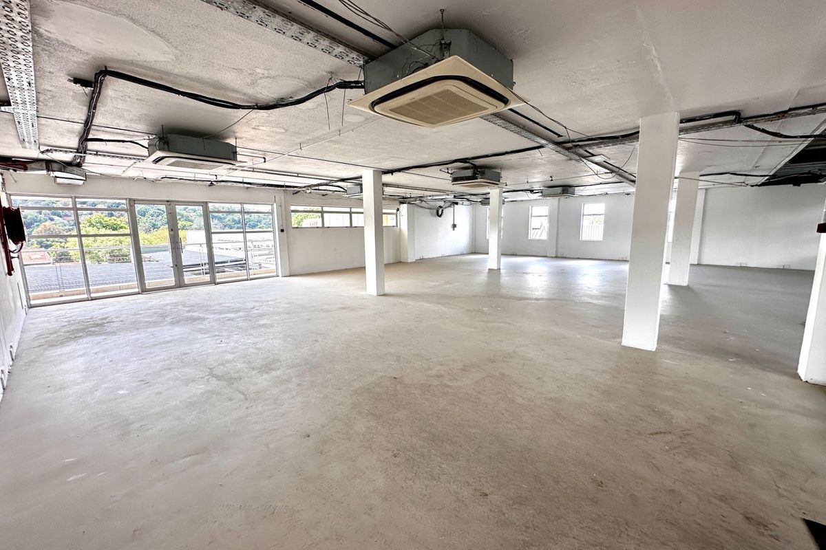 Spacious open-plan office area featuring exposed industrial ceilings, structural columns, concrete flooring, and large glass doors providing natural light.