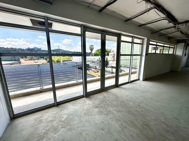 An open-plan office space in shell condition, featuring large glass sliding doors leading to a balcony with expansive views and exposed ceiling infrastructure.