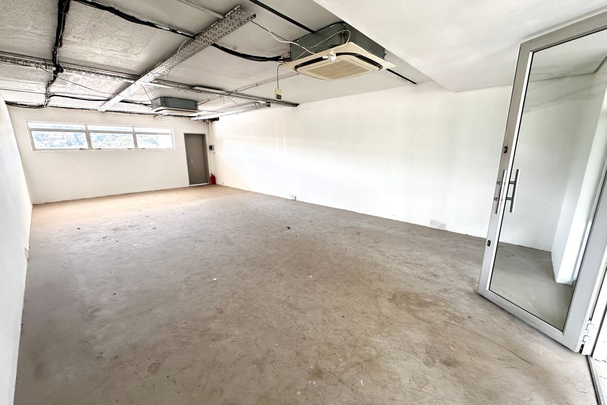 An empty, open-plan office space featuring an exposed ceiling with ducted air conditioning, concrete flooring, and a modern glass entrance door.
